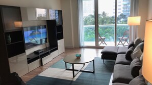 En moderne stue med et fladskærms-tv, en sofa, et sofabord og en balkon med udsigt til palmer og bygninger.
