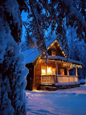 A snow-covered cabin with a wooden exterior, a porch with railings, and string lights. - Cozy, Secluded, Year-Round Cabin: With A Hot Tub! (Munising)