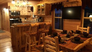 Interno di una baita in legno con cucina, zona pranzo e televisore montato a parete.