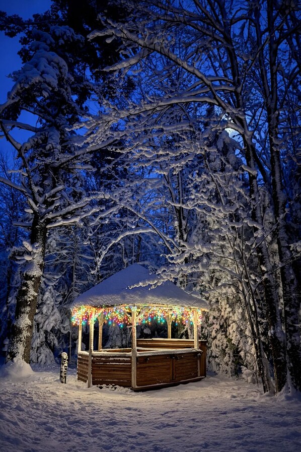 Un pabellón de madera con tejado cubierto de nieve y luces de colores, rodeado de árboles cargados de nieve.