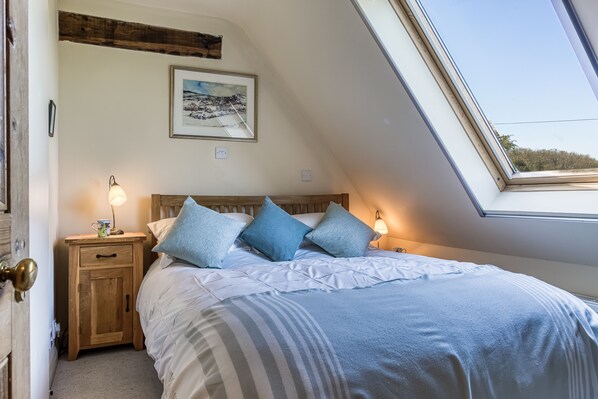 A bedroom with a sloped ceiling, a bed with blue bedding, a nightstand with a lamp, and a framed picture on the wall. - Sheepscombe Byre, Snowshill, Broadway, sleeps 4 guests. This property boasts stunning views. (Broadway)