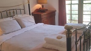Un lit bien fait, avec du linge de lit blanc, une table de chevet en bois sur laquelle se trouve une lampe, et une fenêtre donnant sur un paysage verdoyant.