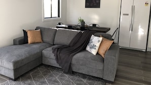A modern living room with a grey sectional sofa, a dining table, and a refrigerator.