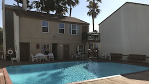 A swimming pool with a wooden deck, a covered patio with outdoor seating, and a two-story building with windows and a balcony.
