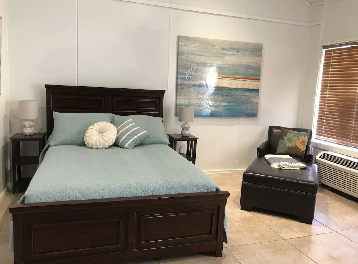 A bedroom with a large bed, a side table, a chair, a small ottoman, and a window with blinds.