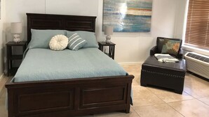 A bedroom with a large bed, a side table, a chair, a small ottoman, and a window with blinds.