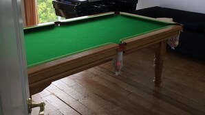 A pool table in a room with wooden flooring, a window with curtains, and a painting on the wall.