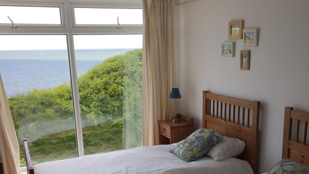 A room with two beds, a window with curtains, a bedside table with a lamp, and framed pictures on the wall.