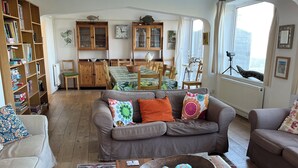 A living room with a grey sofa, a wooden coffee table, and a colorful rug. There is a bookshelf and a dining area in the background.
