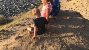 Drie kinderen zitten op een zandstrand met een rotsachtige kustlijn en de oceaan op de achtergrond.