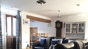 Een woonkamer met een zwarte leren bank, een eettafel met stoelen, een houten kast en een klok aan de muur.