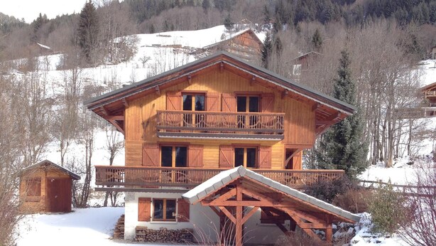 A wooden chalet with a balcony, a snow-covered landscape, and a forested hill in the background.