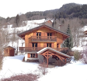 A wooden chalet with a balcony, a snow-covered landscape, and a forested hill in the background.