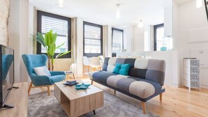 Ein modernes Wohnzimmer mit einer Couch, Sesseln, einem Couchtisch und einem Fernseher. Große Fenster mit Jalousien, eine Pflanze und ein kleiner Beistelltisch sind ebenfalls vorhanden.