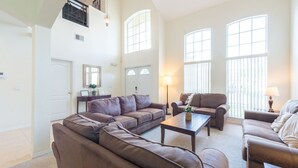 Amplia sala de estar con un sofá modular, una mesa de centro y dos sillones. Grandes ventanas ofrecen luz natural y vistas al exterior.