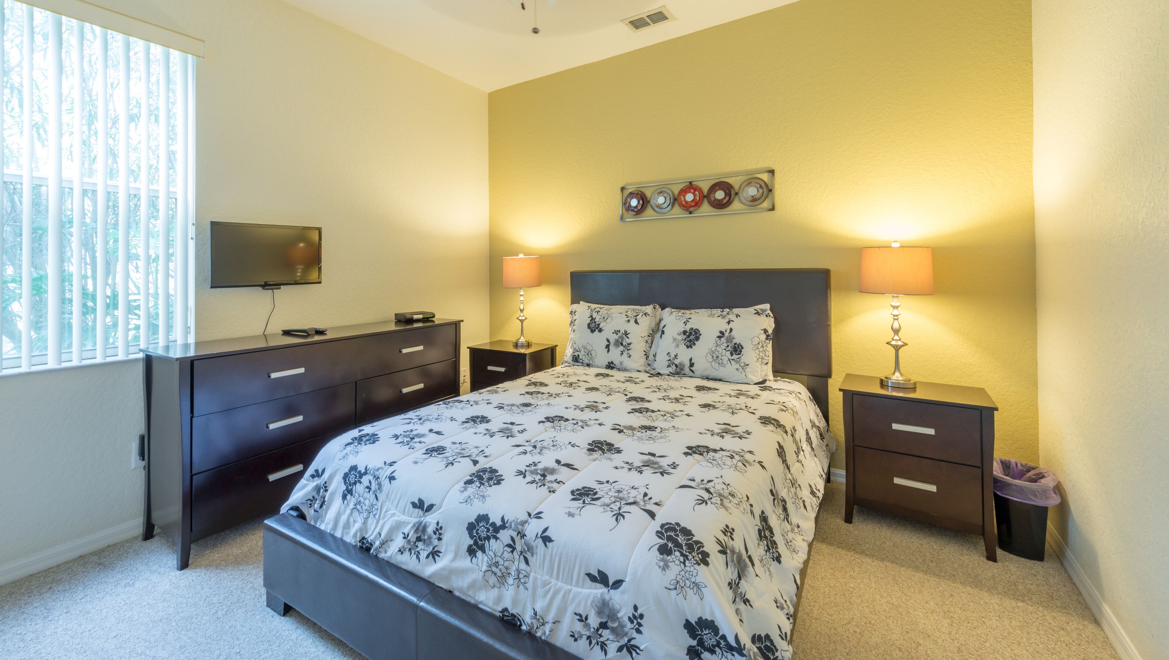 A bedroom with a bed, nightstands, a dresser, a TV, and a wall-mounted light fixture.