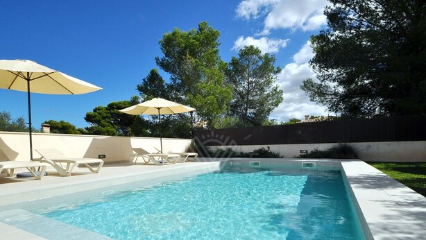 Una piscina rectangular con agua cristalina, rodeada de sillas de descanso blancas y dos sombrillas color beige.