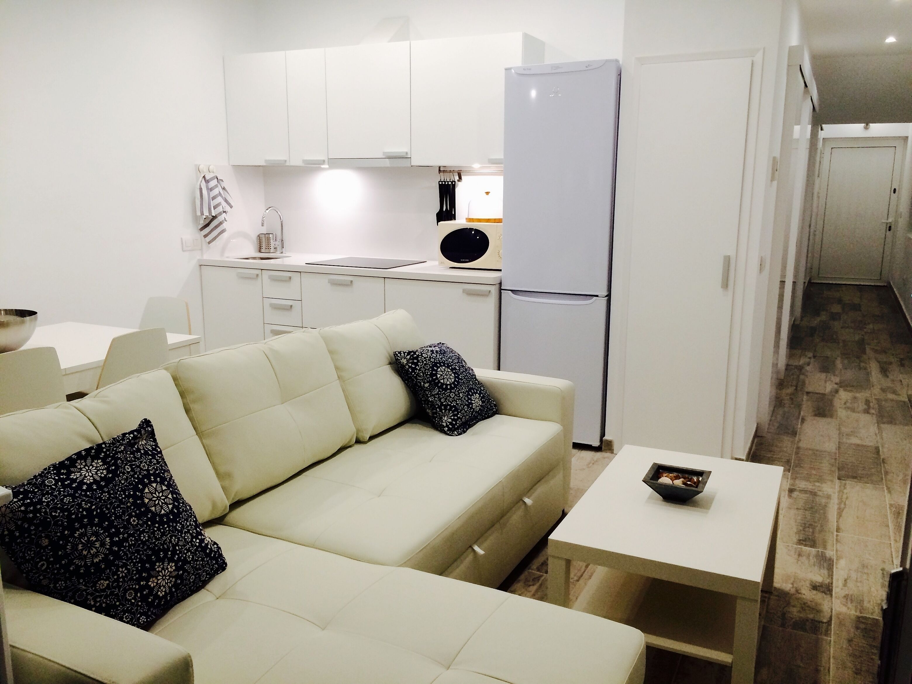 Ett kompakt bostadsutrymme med en soffa, ett litet bord och ett köksområde som inkluderar ett kylskåp och skåp.