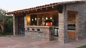 Une cuisine extérieure avec une façade en pierre, un four à pizza, des tabourets de bar et un barbecue.