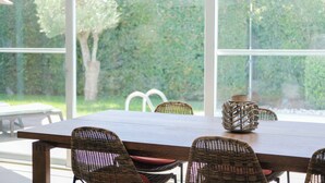 Un comedor con una mesa de madera y sillas de mimbre, amplias ventanas con vistas a un seto verde y árboles.