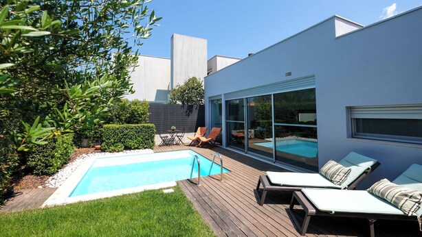 Un área de piscina moderna al aire libre con muelle de madera, sillones de descanso y un edificio con paredes de cristal.