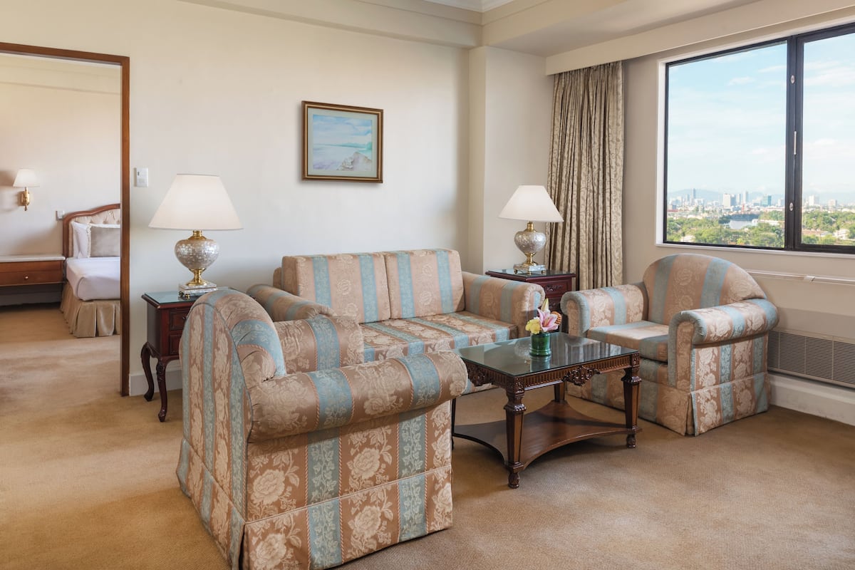 A hotel room with a sofa set, two lamps, a painting, and a large window with a city view.