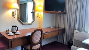 Habitación de hotel con escritorio de madera, una silla, televisor, espejo, ventana con cortinas, radiador y cama.