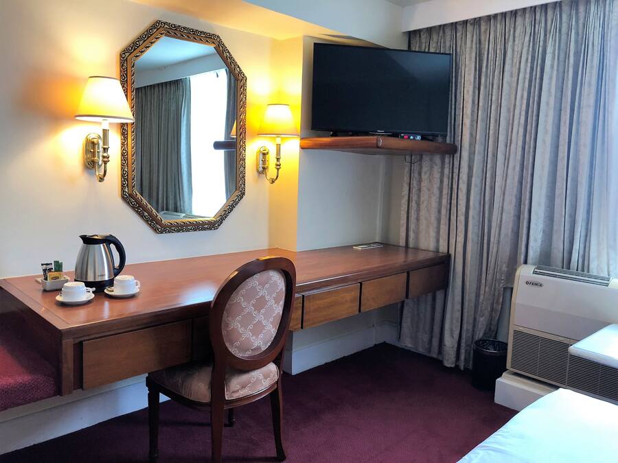 Habitación de hotel con escritorio de madera, una silla, televisor, espejo, ventana con cortinas, radiador y cama.