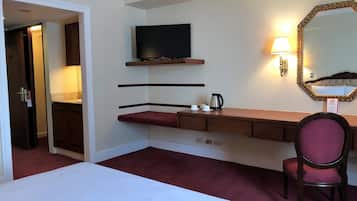 Ein Hotelzimmer mit einem Bett, einem Schreibtisch mit Stuhl, einem Fernseher, einem Spiegel und einem an der Wand befestigten Ablagefach mit Wasserkocher.
