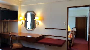 Habitación de hotel con escritorio, silla, banco, televisor, espejo, apliques de pared y una puerta que da a otra habitación.