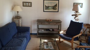 Un soggiorno con un divano blu, un tavolino in legno con quotidiani, un tavolino laterale anch'esso in legno con una lampada, e una poltrona.