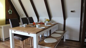 Een eetruimte met een houten tafel, witte stoelen en bankjes. Er staat een kleine tafel met een fruitschaal en kopjes. De kamer heeft houten balken in het plafond en een witte deur.
