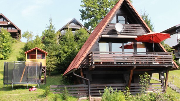 Een houten chalet met een balkon en een rode parasol, omringd door bomen en een speeltuin.