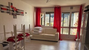 A room with a white sofa, a wooden chair, a bed with a red blanket, and a window with red curtains.