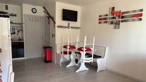 A modern kitchen with a clock, a dining area with white chairs and red cushions, and a TV mounted on the wall.