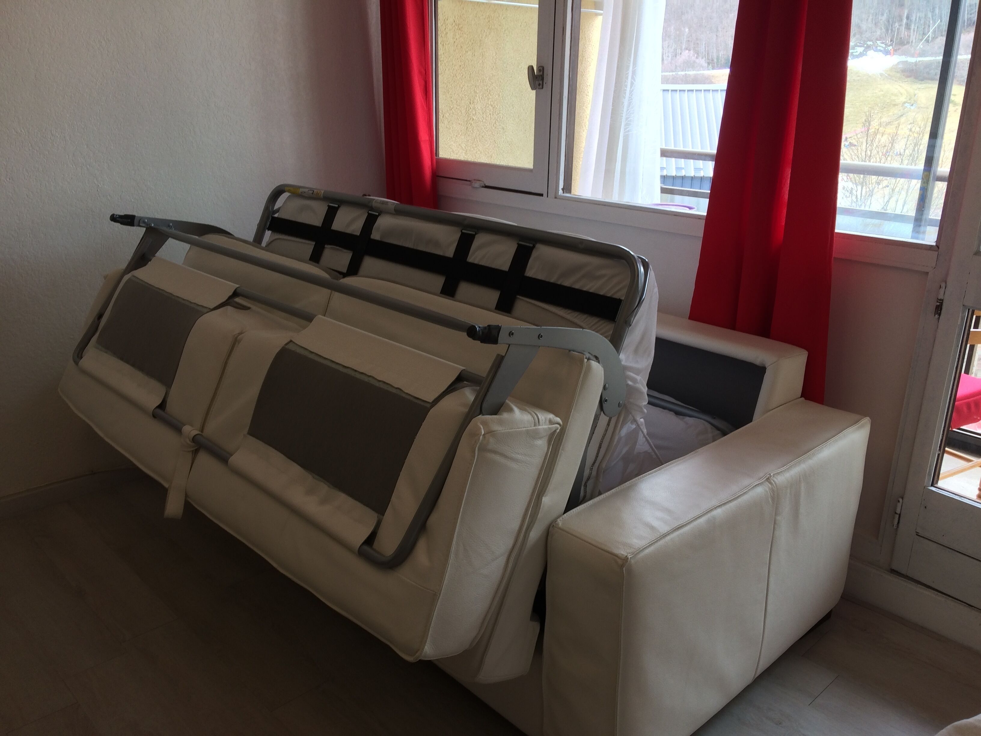 A white sofa with a metal frame and a cushion, positioned near a window with red curtains.