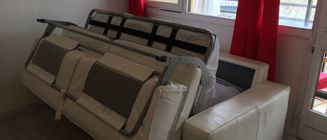 A white sofa with a metal frame and a cushion, positioned near a window with red curtains.