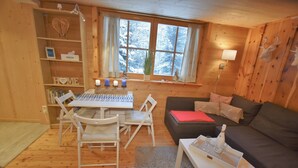 Ein gemütliches Blockhaus mit Holzvertäfelung, einer Essecke mit Tisch und Stühlen, einem Sofa mit Kissen und einem Fenster mit Blick auf schneebedeckte Bäume.