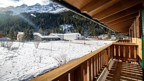A wooden balcony with a view of a snowy landscape, a mountain range, and buildings in the distance.