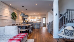 Un intérieur moderne avec un plancher en bois, un coin repas comprenant une table et des chaises, un canapé recouvert d’une couverture à carreaux rouges et blancs, et un escalier avec une rampe en bois.