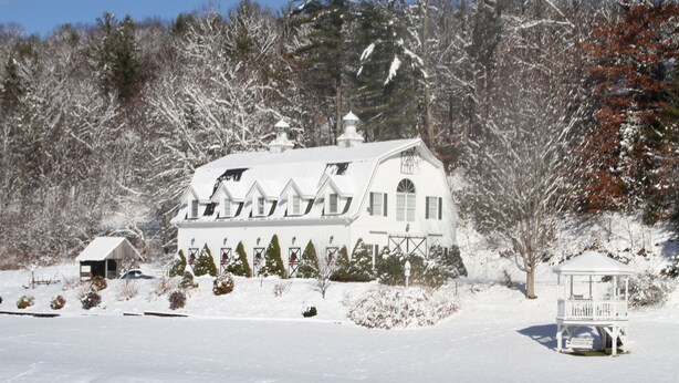 Ein großes, weißes, mehrstöckiges Haus mit symmetrischer Dachlinie und mehreren Schornsteinen, umgeben von schneebedeckten Bäumen und einer kleinen Laube.