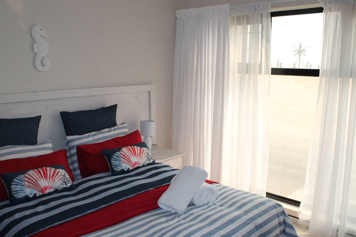 Una cama bien hecha con ropa de cama a rayas y almohadas rojas, una mesita de noche blanca con una lámpara, y vista a una playa a través de cortinas traslúcidas.