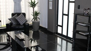 Una sala de estar moderna con un sofá de cuero negro, una mesa de centro de vidrio, una planta grande en maceta y una estantería con diplomas enmarcados.