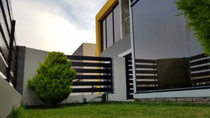 Un edificio moderno con techo plano, ventanas amplias y un pequeño jardín con un arbusto y una manguera.