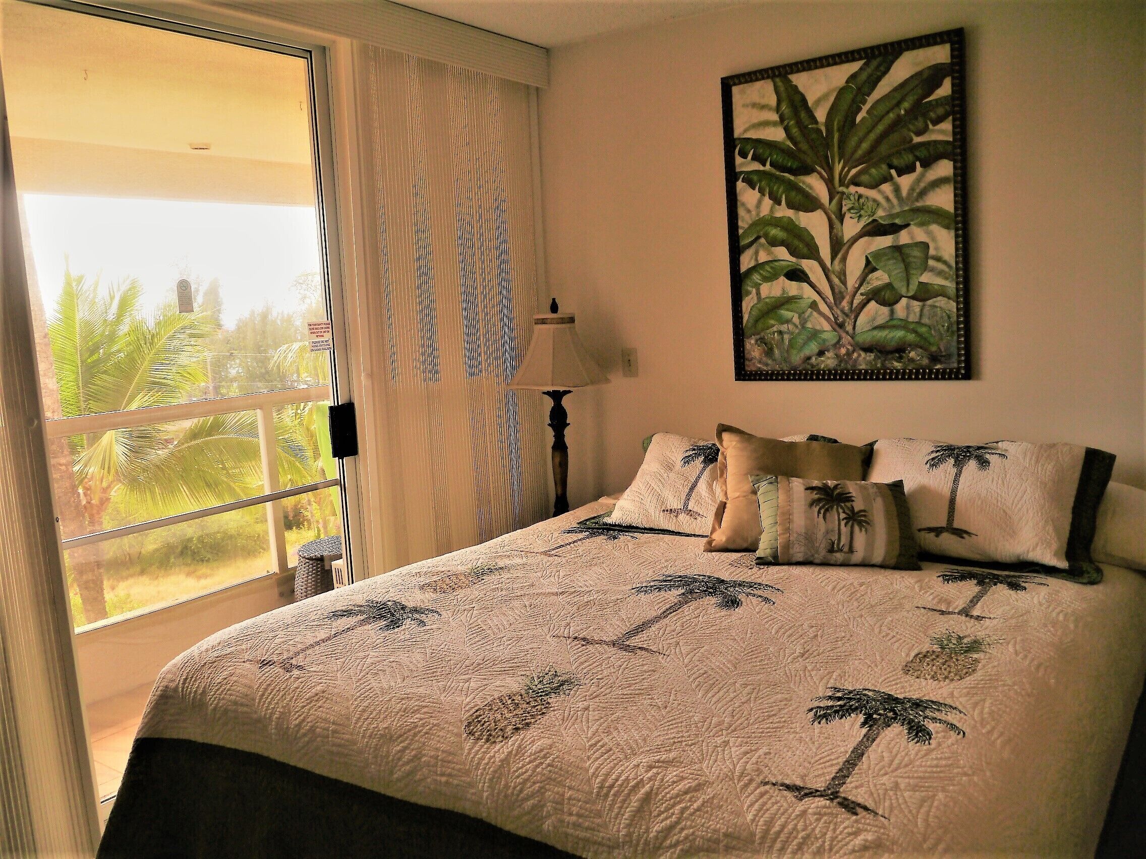 A bedroom with a large bed, a window with curtains, a painting of a plant, and a nightstand with a lamp.