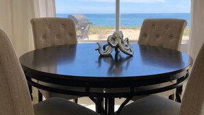 Une table ronde à la surface sombre, entourée de quatre chaises beiges à revêtement rembourré. Une sculpture décorative représentant une pieuvre est posée sur la table. La pièce est dotée de grandes fenêtres ornées de rideaux blancs, offrant une vue sur la plage et l’océan.