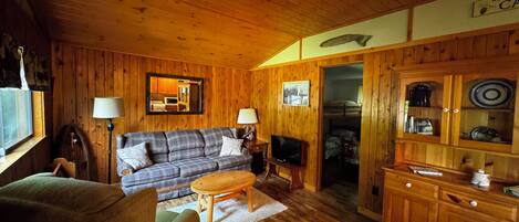 A cozy cabin interior with a wooden ceiling, a sofa, a television, a dining table, and a cabinet with dishes.