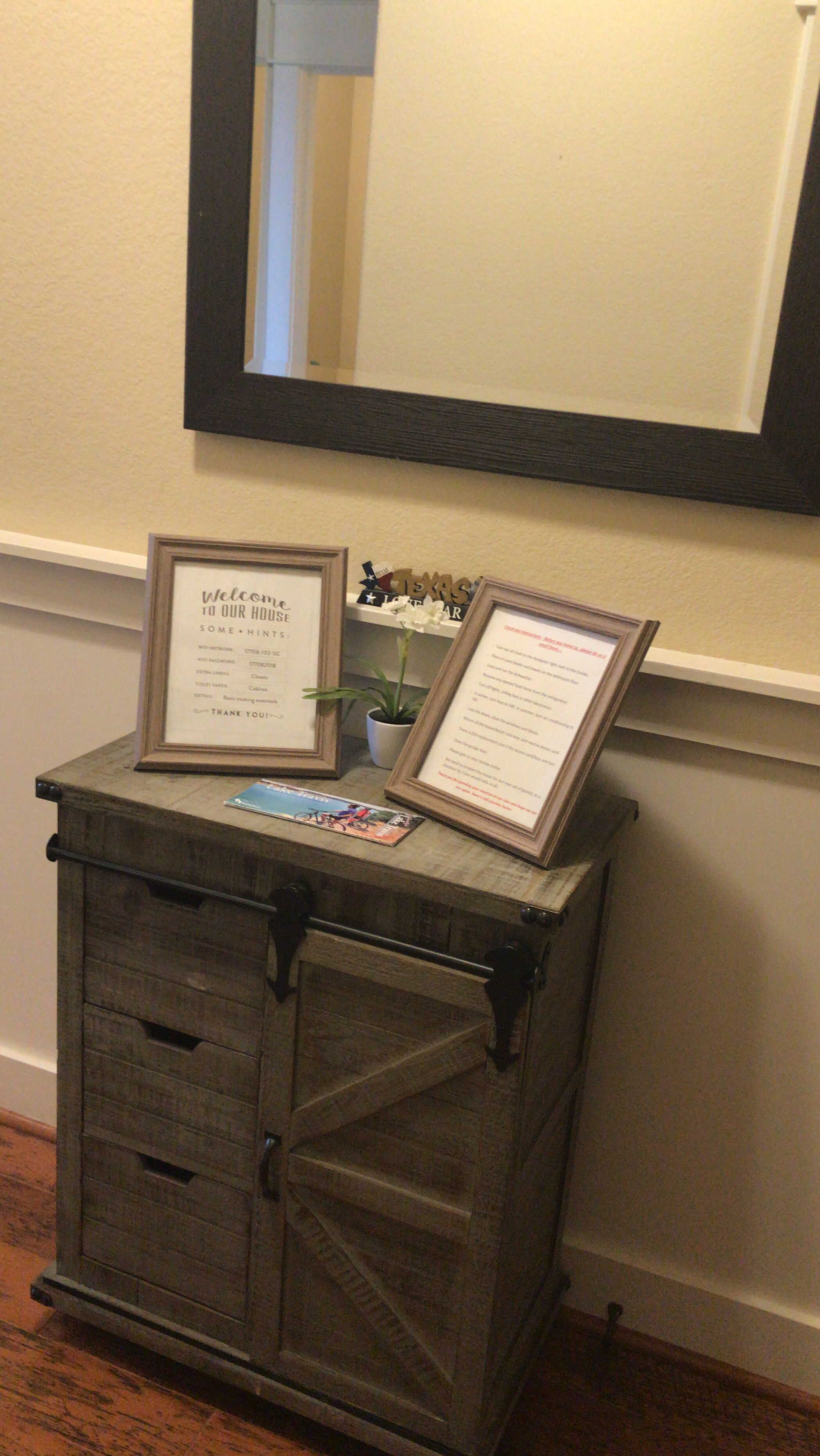 A wooden cabinet with drawers and a mirror above it. There are two framed welcome signs on the cabinet.