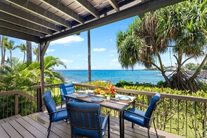 Un área de comedor al aire libre cubierta con una mesa dispuesta para dos, con vistas a una playa con palmeras y el océano.