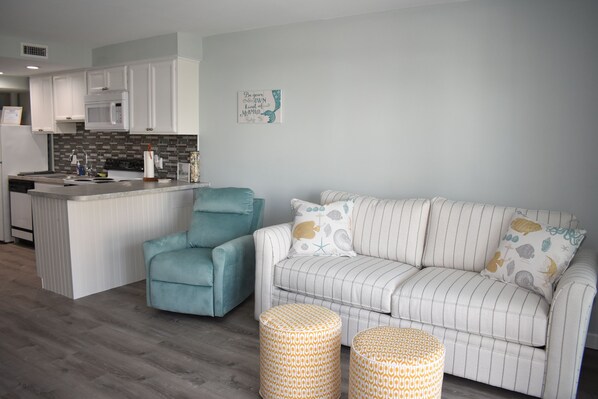 A modern kitchen with white cabinets and a built-in microwave. There is a teal armchair and a striped sofa with decorative pillows. Two patterned ottomans are placed in front of the sofa.